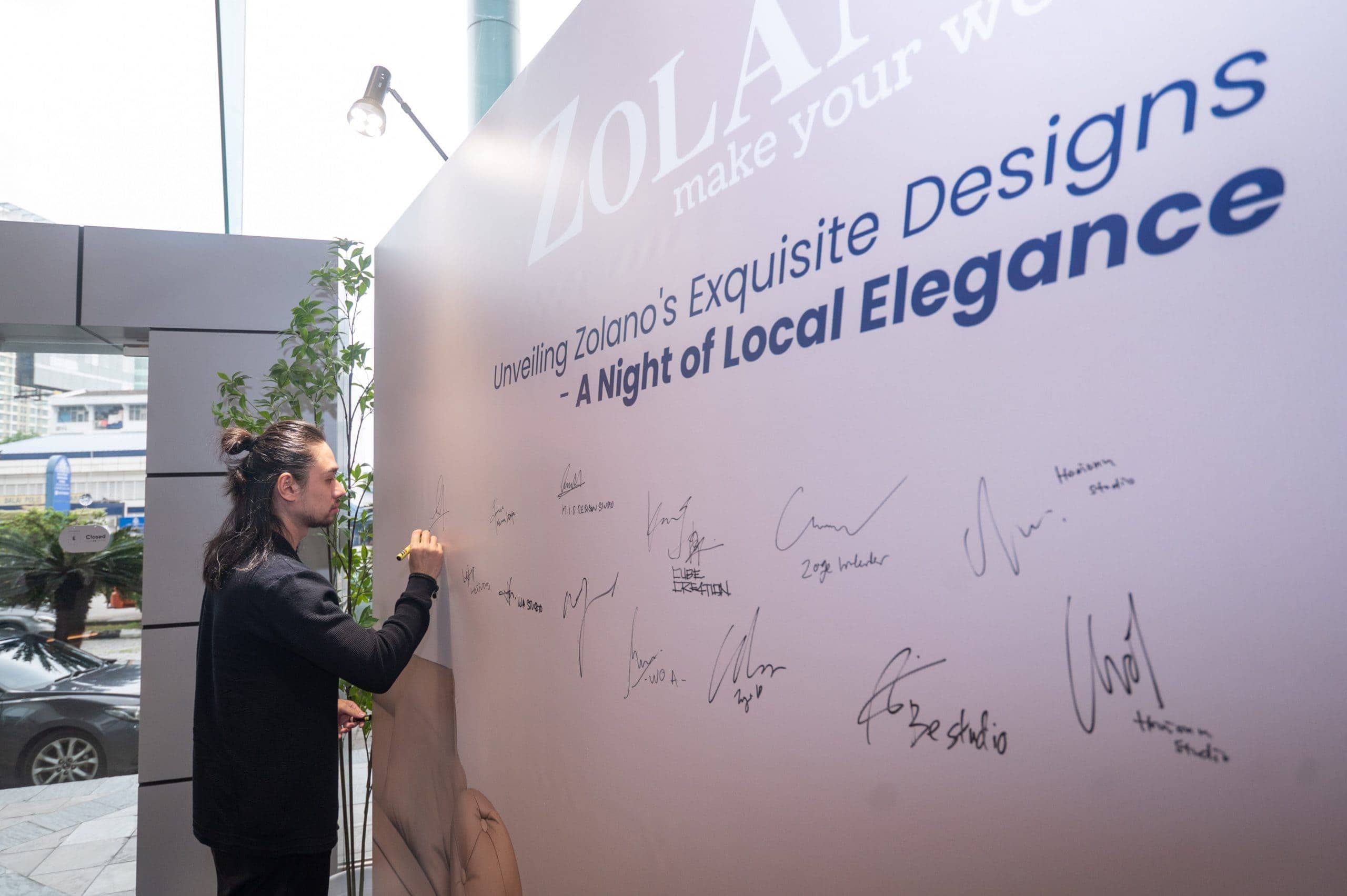 Elegant woman signing a Zolano event wall with furniture collection unveiling signage.