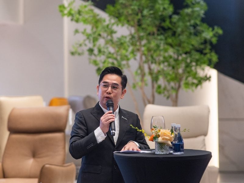 A man speaking at a furniture event with elegant sofas and greenery in the background.